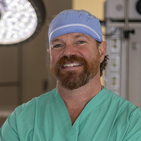 A surgeon in green scrubs and a surgical hat stands in an operating room