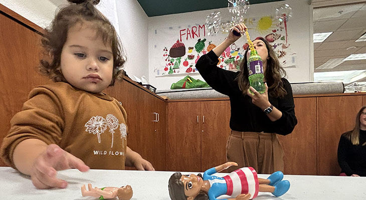 a young girl plays with plastic toys at a table while a woman blows bubbles in the background