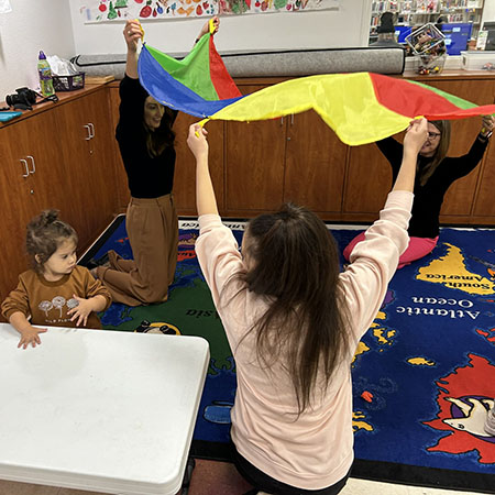 Three women hold a colorful parachute over their heads while a young girl plays nearby.