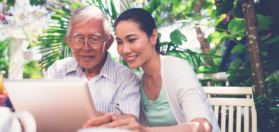 Older man sits next to daughter outdoors while both look at laptop screen in front of them