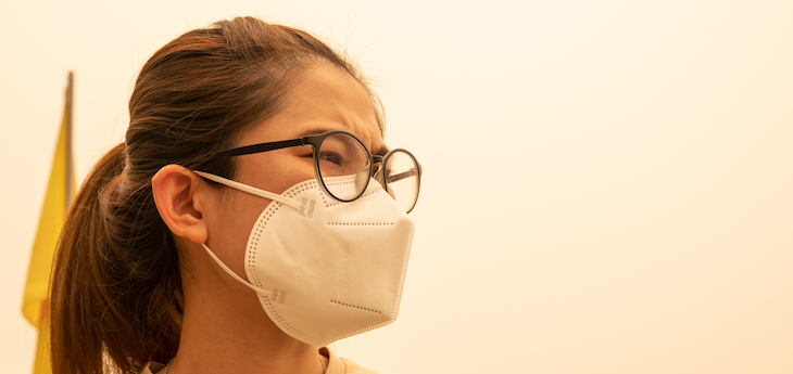 Worried-looking woman wears a mask against a hazy pink background of bad air.