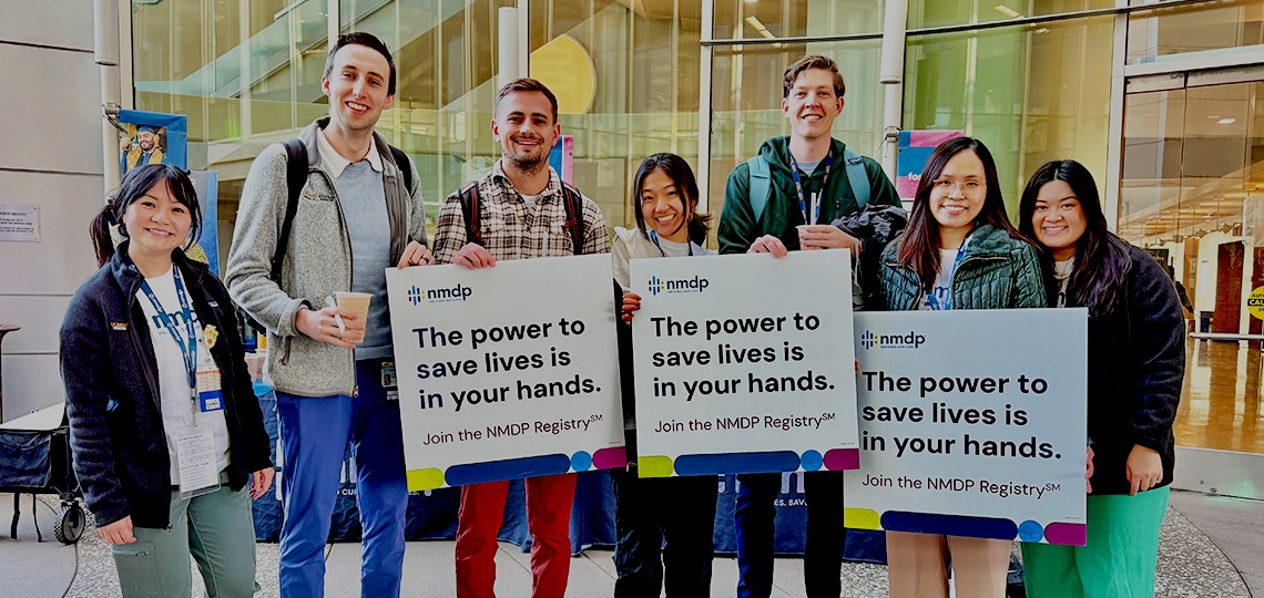 Seven medical students stand in front a building while holding signs encouraging signups for the National Marrow Donor Program