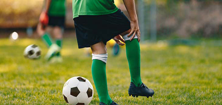 A boy wearing a green and black soccer uniform reaches down to hold his knee, a soccer ball next to him on a field.  