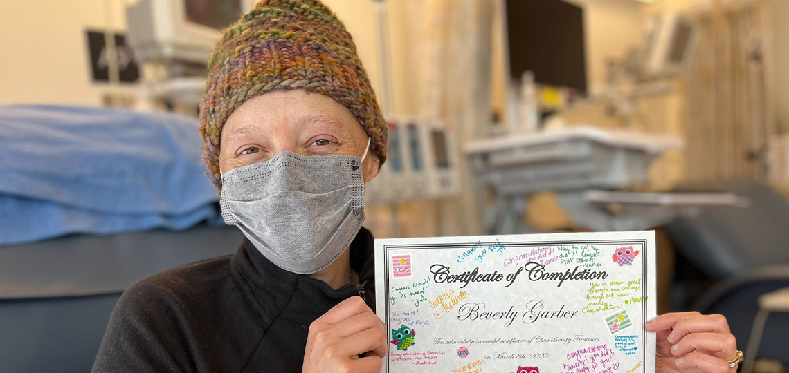 Woman with a tan knit hat and wrapped in a blanket and holding a certificate.