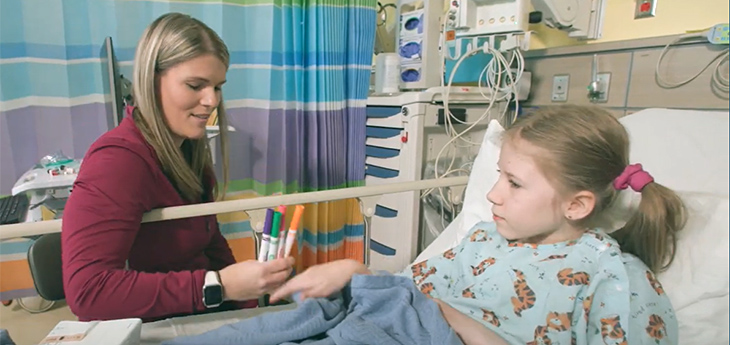 Woman holds markers as child lays in hospital bed smiling