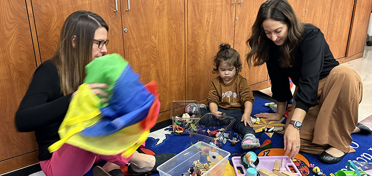 Two woman and a young girl play with colorful toys while seated on a colorful rug at a library playgroup