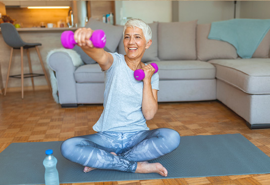 Woman sits with crossed leg on the floor on a yoga mat holding weights with one arm extended out