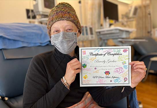 Woman with a tan knit hat and wrapped in a blanket and holding a certificate.