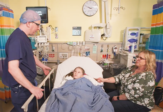 Doctor looks over patient in hospital bed, mom sits next to her.