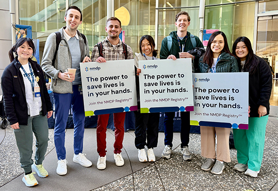 Seven medical students stand in front a building while holding signs encouraging signups for the bone marrow registry program