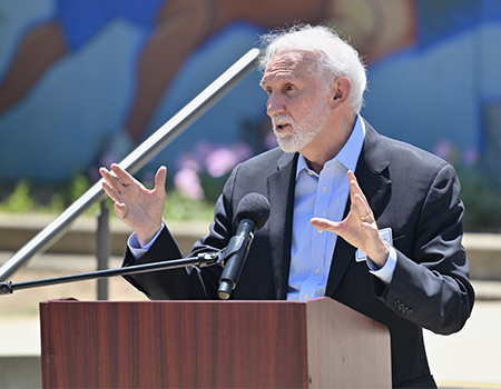 A man with a white beard in a navy blue suit jacket and blue collared dress shirt gestures with both hands at a podium outdoor