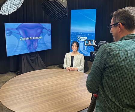 A woman sits behind a table for a television interview, with a TV screen to her right that says 