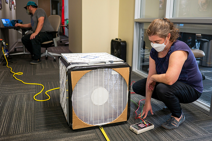 Woman in mask kneels next to a Corsi-Rosenthal box.