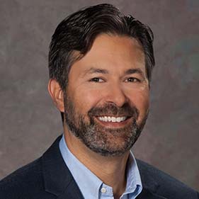  A man in a dark blue suit and light blue shirt with dark brown hair and a beard is smiling.