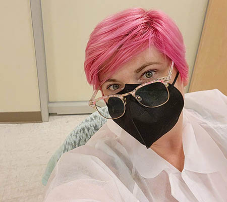 A researcher with pink hair sits at a desk wearing a black mask.