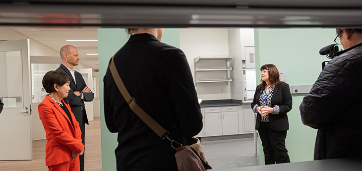 A woman in a dark suit stands in a research lab with green walls while speaking with a man and a woman to her right