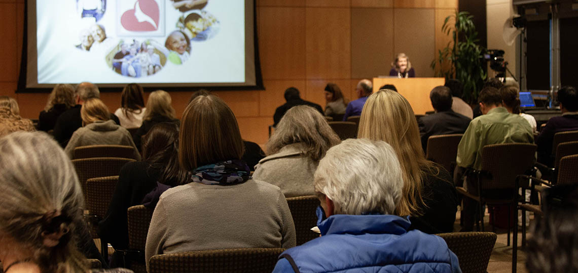 A crowd of seated people look toward a podium where a woman gives a lecture in an auditorium.