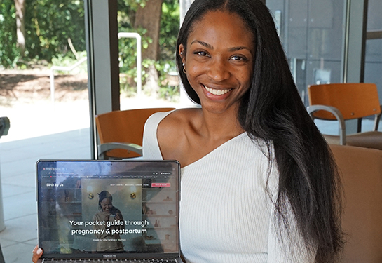 A woman wearing white sweater holds laptop with its screen that reads: “Your pocket guide through pregnancy and postpartum”