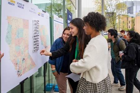 Three women stand before a poster-sized map of California that shows pin drops of where UC Davis students will train for residency