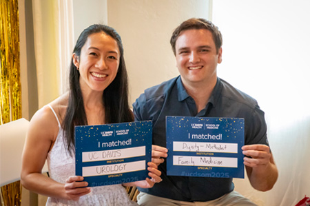 A woman in white dress and man in blue shirt sit on chairs holding signs announcing they matched at UC Davis and Dignity Methodist