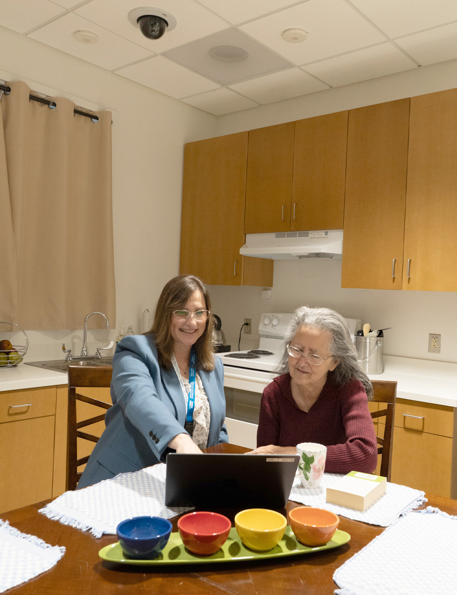 Professor Fritz, left, sits next to older adult woman at kitchen table pointing at laptop in front of them