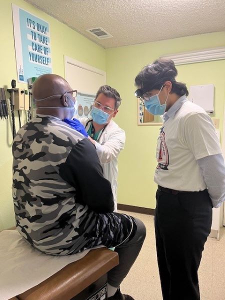 Back of male patient sitting on exam table with two people talking to him.