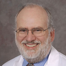 A physician in a white coat with a gray beard, wearing glasses