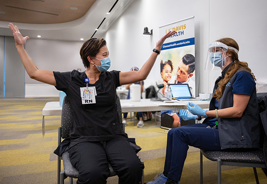 A nurse in blue scrubs raises her hands in jubilation after receiving a COVID-19 vaccine