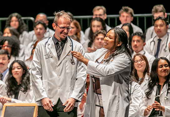A new medical student in white coat smiles as a faculty member places a stethoscope around his neck