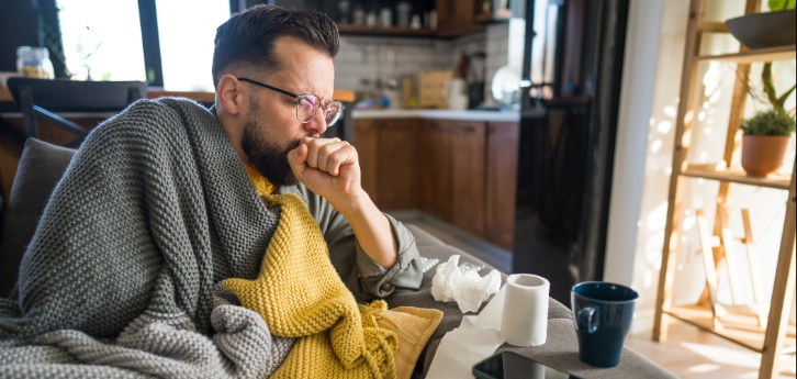 Sick man on sofa coughing, wrapped in blanket, surrounded by tissues and toilet paper, feeling unwell.