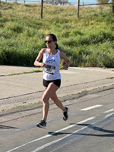 Woman in sunglasses, white tank top with a numbered bib and black shorts runs along a paved road