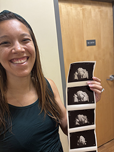 Smiling woman in front of a brown door holding a photo strip of 3-D ultrasound images of her developing baby.