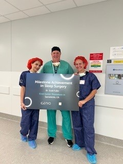 Three people in surgical attire stand in a hallway holding a sign that reads