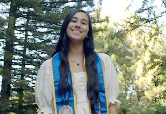 Smiling woman outdoors wearing a light dress and blue academic stole.