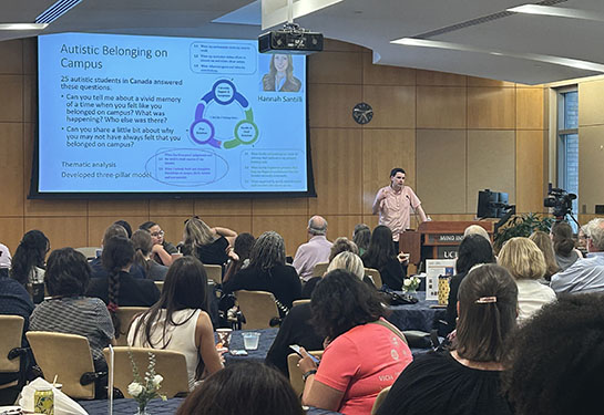 A speaker presents to a crowd in an auditorium with a large screen behind them.