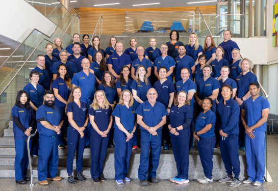 Dozens of emergency medicine team members in matching blue scrubs stand on a staircase inside a modern building.