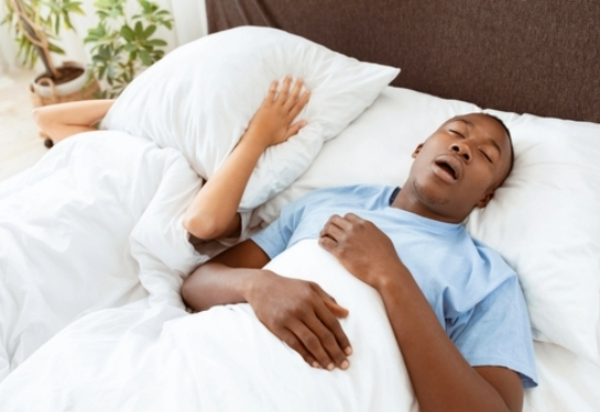 Two people are lying in bed, with one person resting under a white blanket and the other partially covered by a pillow.