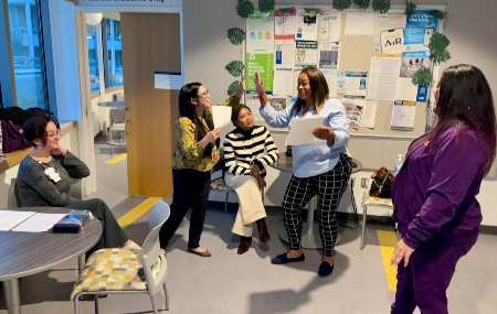 Employees gather in a medical student lounge for a team activity, with one person speaking while colleagues listen and participate.