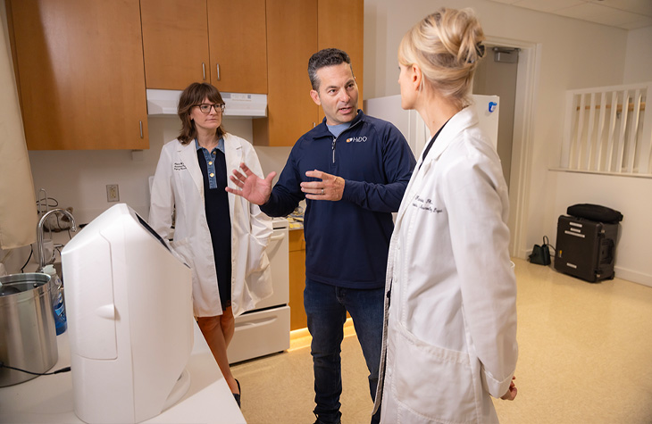 Three people discuss a smart medication dispenser in a simulated kitchen space.