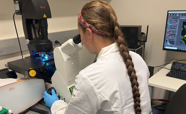 A researcher in a white lab coat is seated behind a microscope station, leaning over to examine something. 