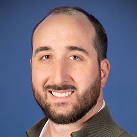 Thomas Ambrosi, in a brown collared jacket and white shirt, smiles. 