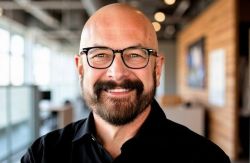 Portrait of Chris Long wearing glasses and a black shirt in an indoor setting.