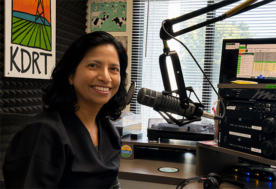 Ulfat Shaikh smiles sitting in front of a microphone in a studio.