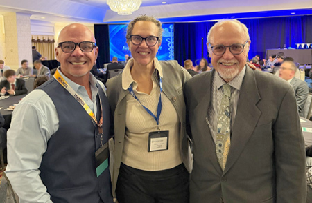 Three smiling professionals wearing conference badges pose together at an event, with attendeesat tables and a stage in the background.