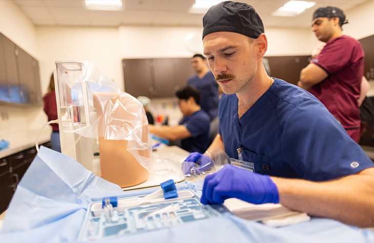 Male nursing student wearing blue scrubs and purple gloves practices a clinical procedure in a simulation lab.