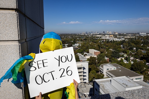 A person dressed up in a blue and yellow bird costume holds sign that reads, see you Oct. 26
