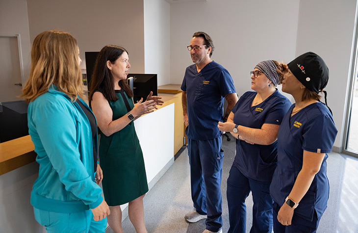 A woman in a green dress speaks with four healthcare professionals, three wearing navy blue scrubs