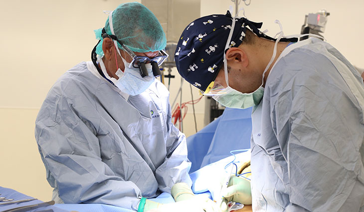 Two surgeons in blue scrubs operate on a patient in an operating room.