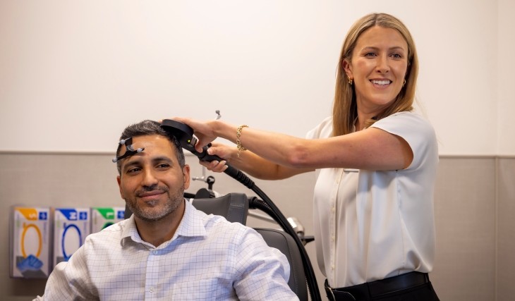 A woman standing in a medical setting holds a TMS therapy device over the head of a seated man.