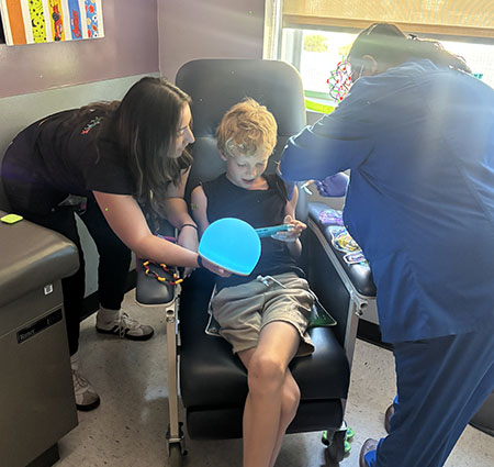 A young boy sits in a medical chair while several adults around him prep him for a vaccine